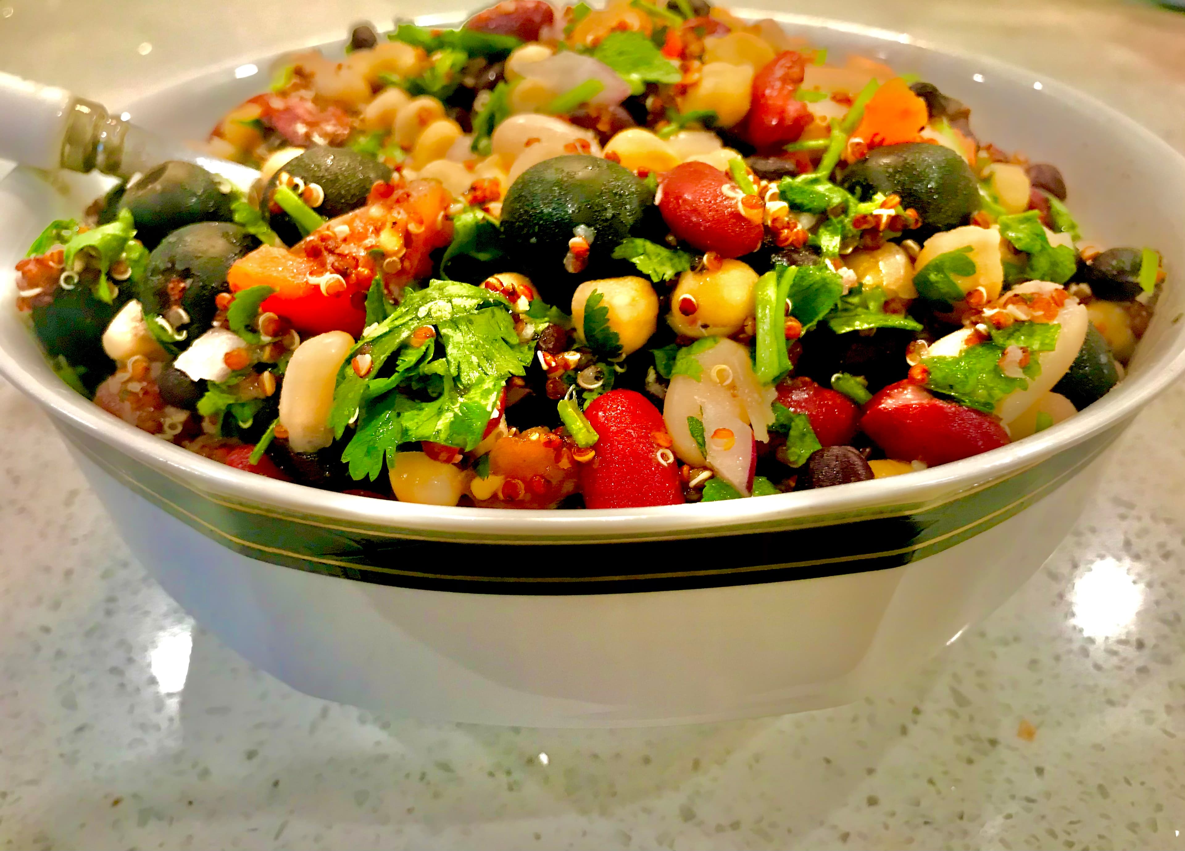A bowl of legume salad with beans, lentils, quinoa, tomatoes, olives, and cilantro